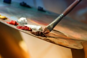 An artist dipping their paintbrush in brown acrylic paint on an easel.